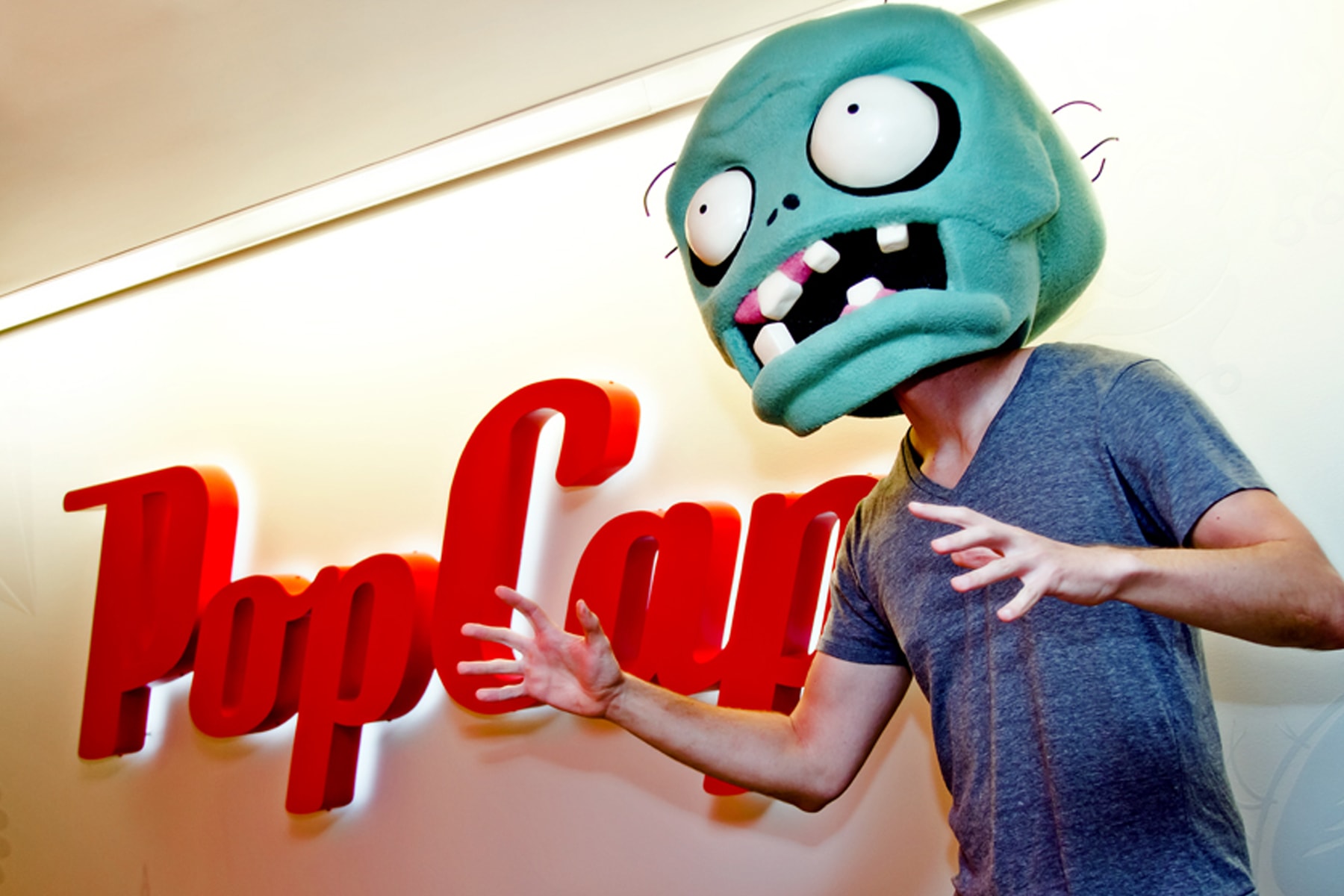 Mark Barrett wearing the zombie mascot's head in front of a lit, wall-mounted PopCap logo