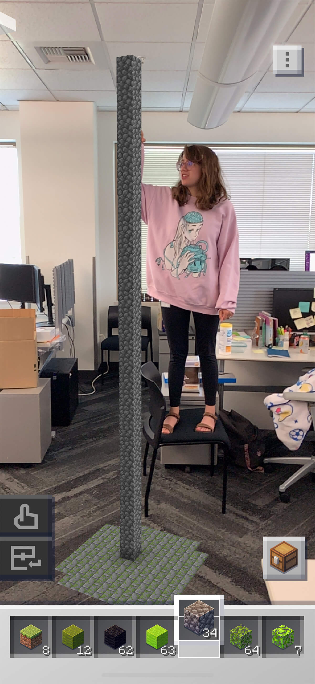 DigiPen graduate and Mojang artist Kelly Greene stands on a chair in her office next to a tall augmented reality Minecraft Earth structure