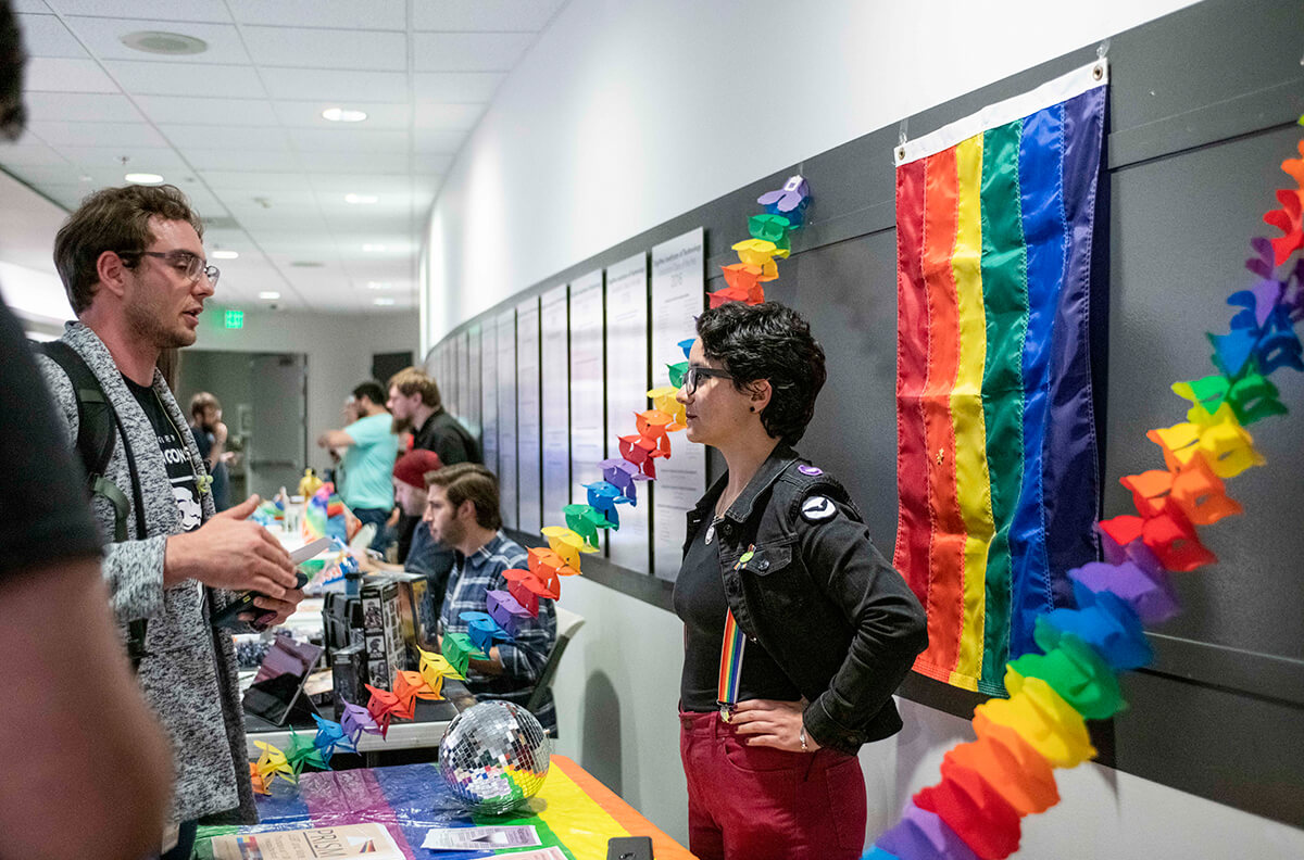 Students converse at a club info event; PRISM club representative stands at a table draped with rainbow-colored tablecloth and accordion streamers.