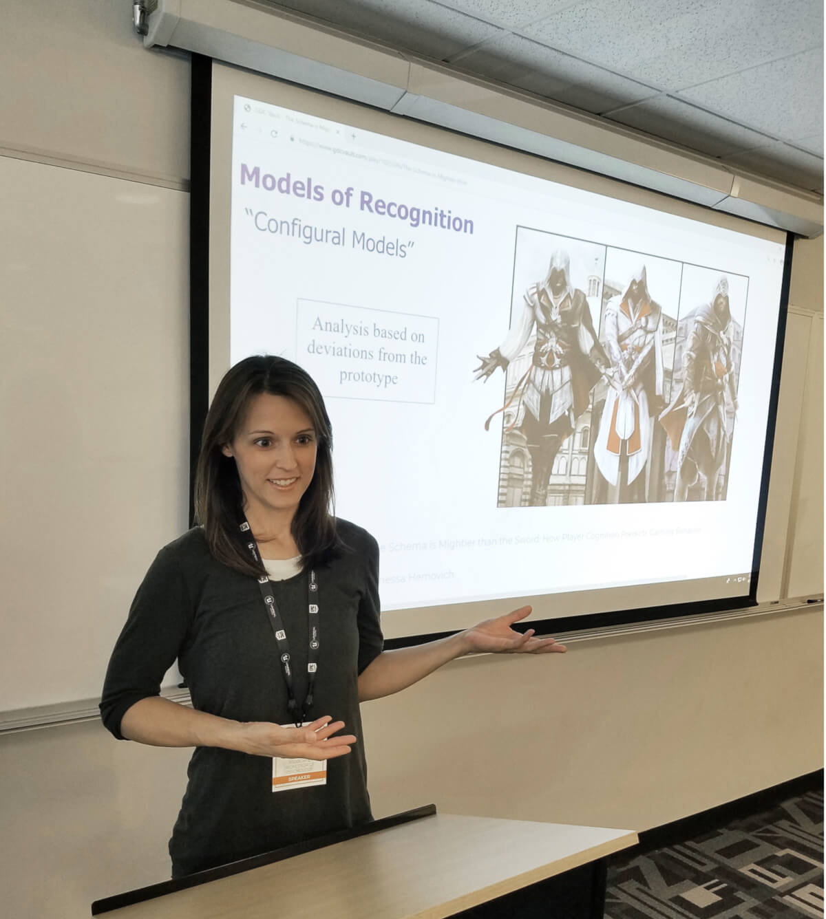 Professor Vanessa Hemovich lectures while gesturing at a slide showing characters from the game Assassin's Creed.