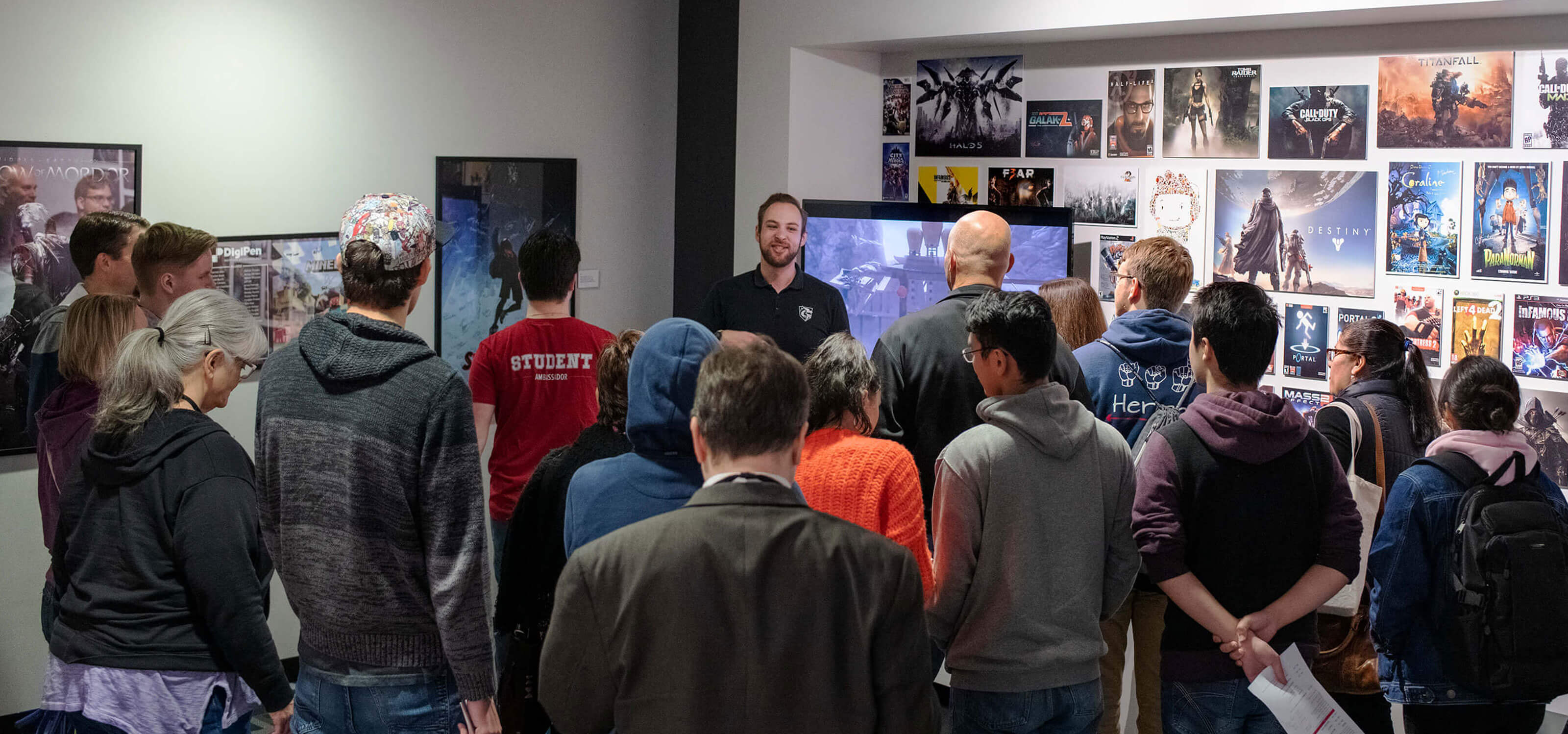 A group of people seen from behind as a man in a black polo shirt addresses them in a hallway strewn with video game posters.