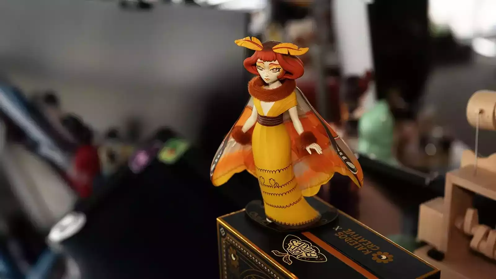 A close up of the Atlas figurine, a moth fairy wearing a yellow dress, posed atop its black box packaging. 