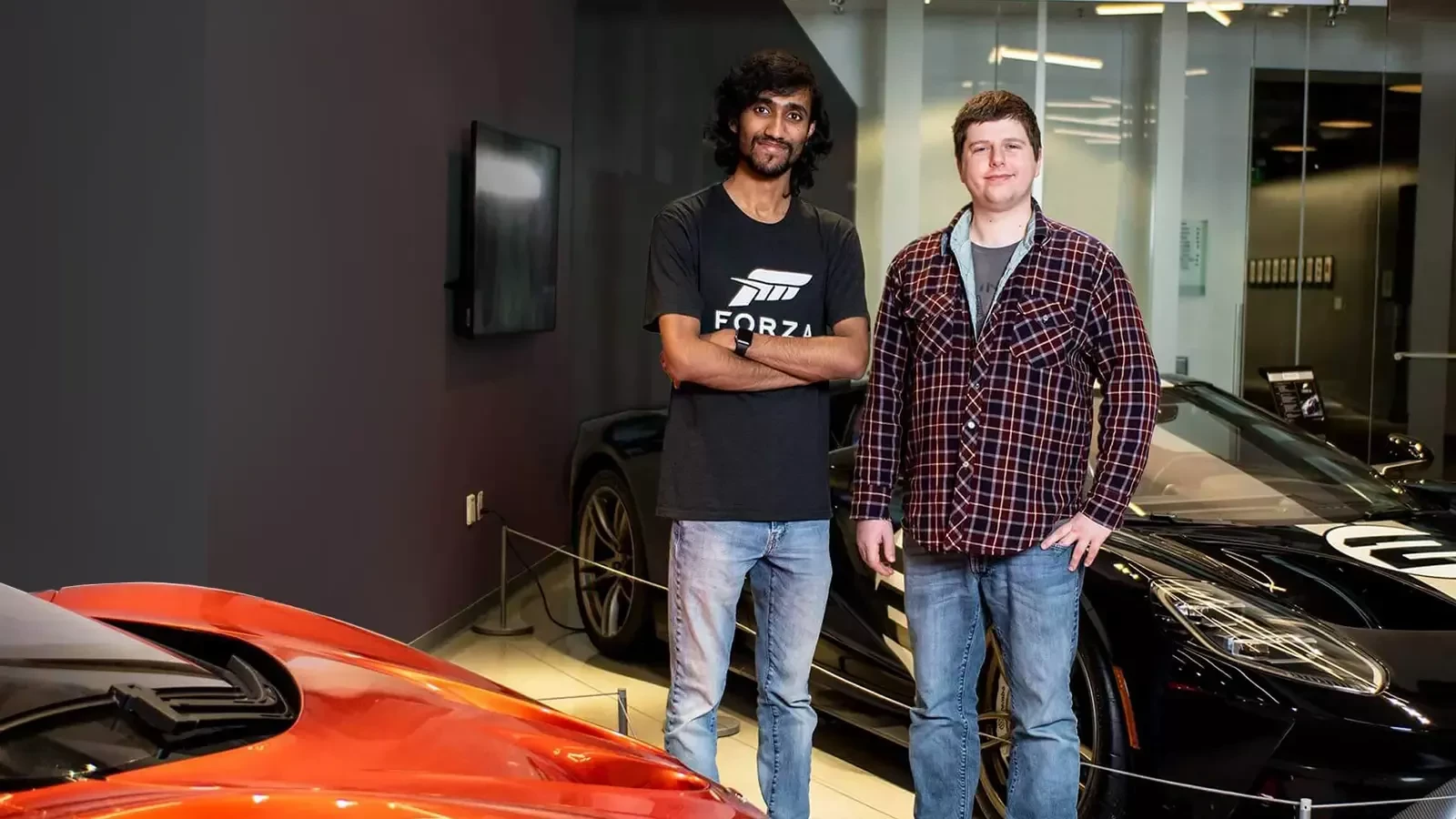 Two alums stand next to luxury cars in the Forza game studio lobby.