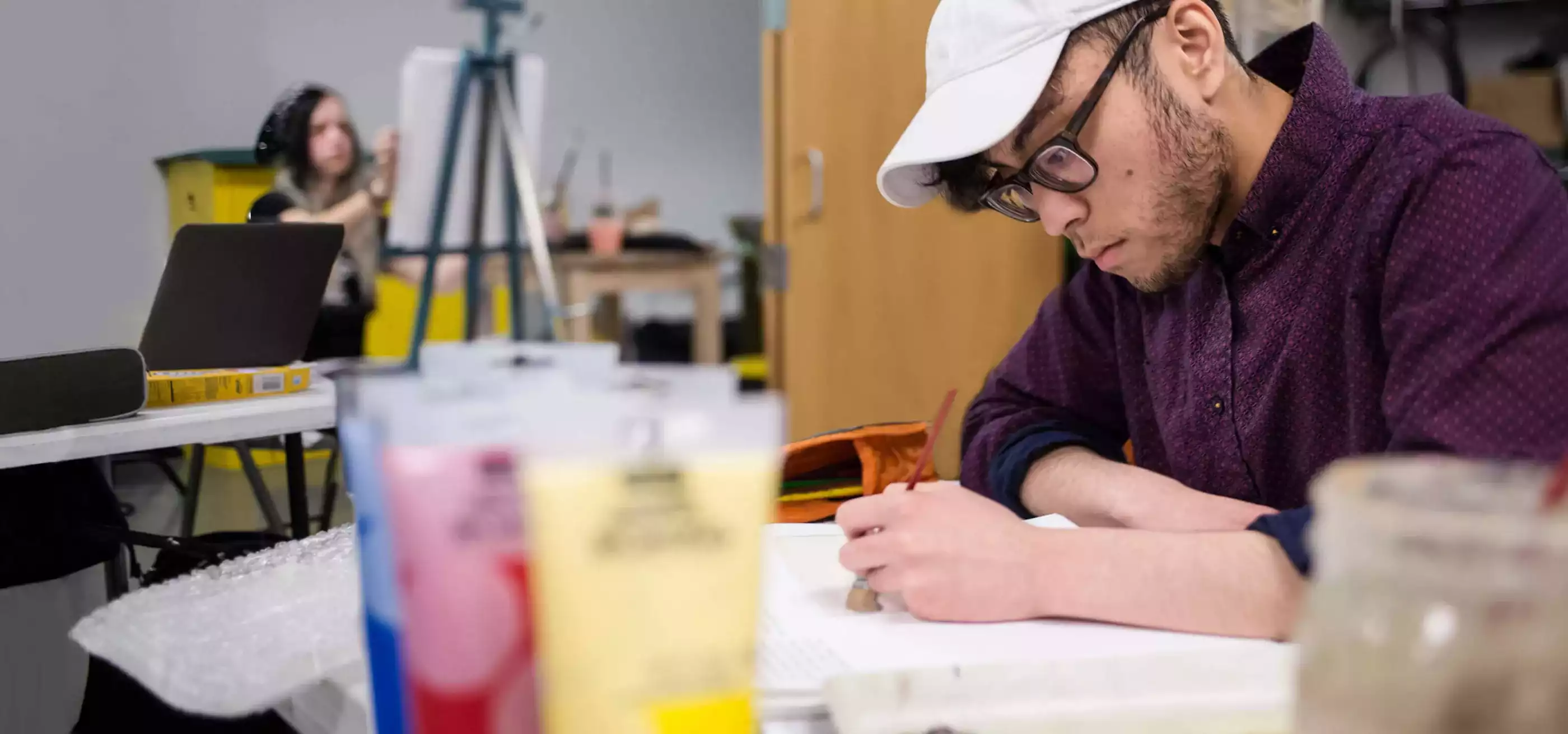 A man looks intently at the paint he is applying to a piece of paper. A woman in the background is painting with an easel.