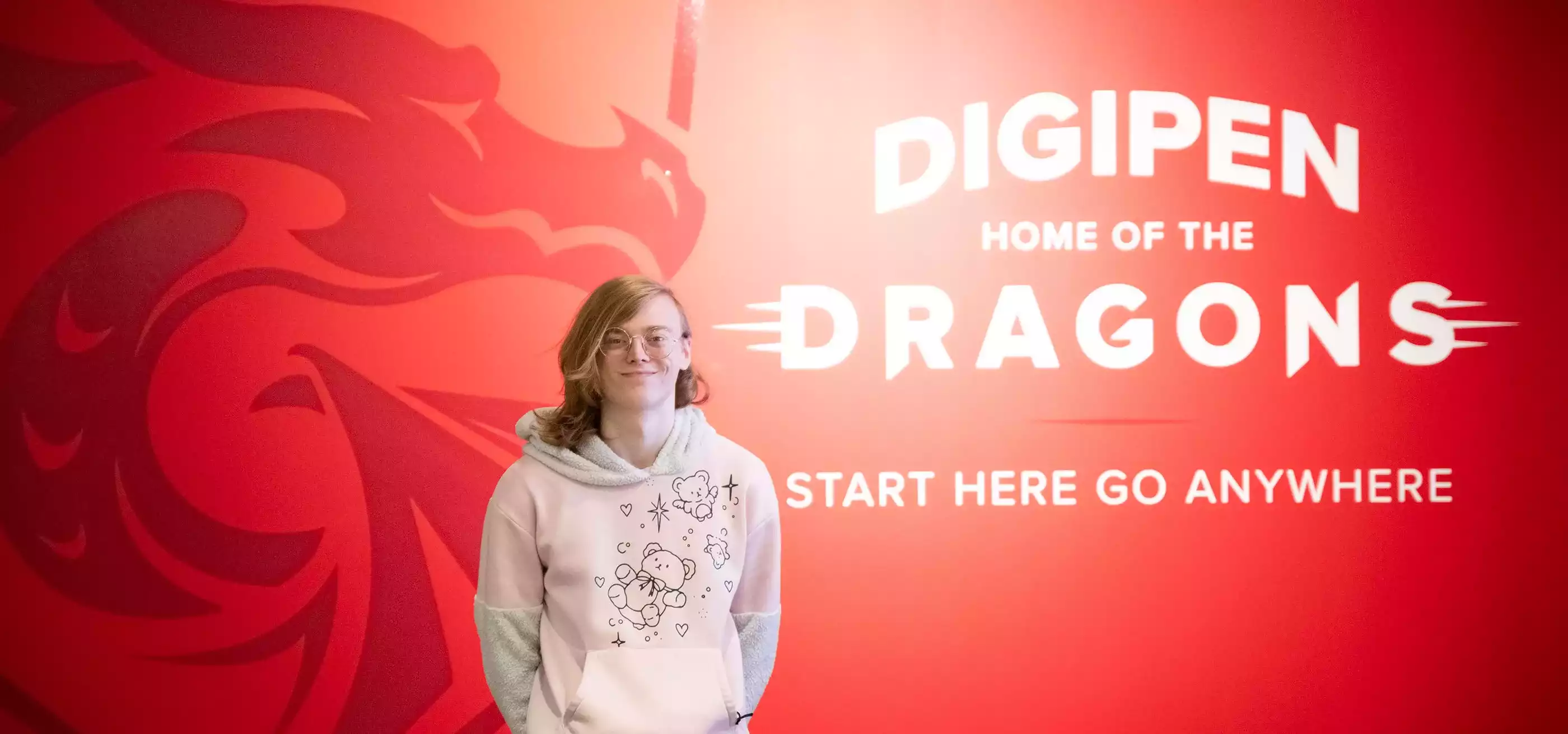 Student Tetra Chaney poses in front of a red wall with DigiPen’s Dragon logo on it and the words, “DigiPen Home of the Dragons — Start Here Go Anywhere.”