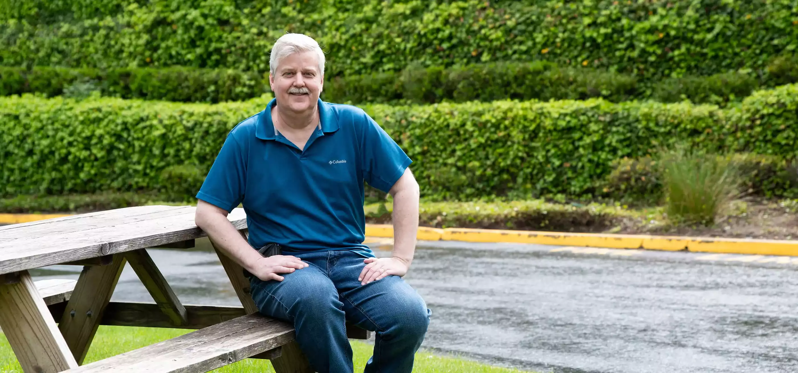 Douglas Schilling sits at a picnic table outdoors with his hands on his knees and smiles.