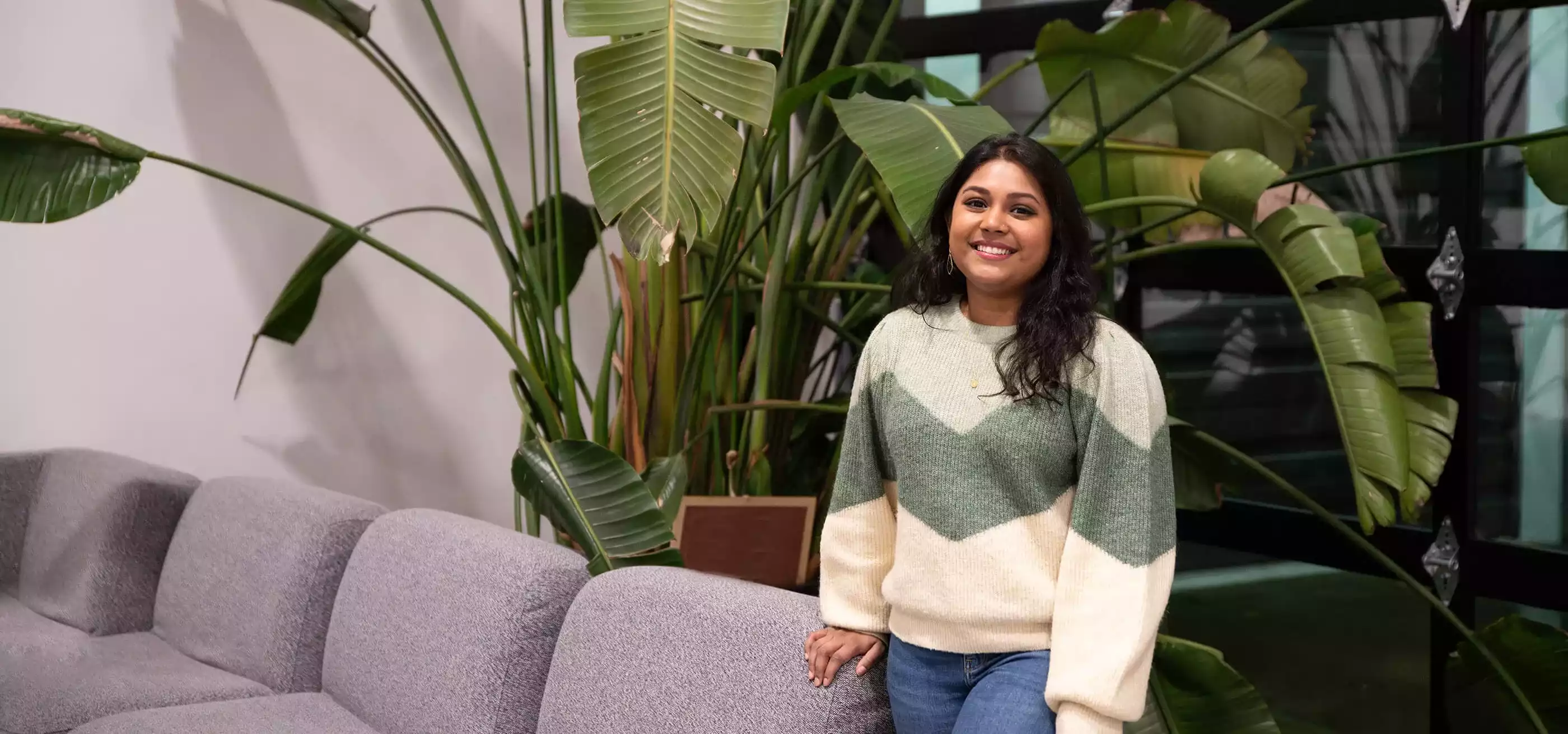 DigiPen graduate Neha Chintala smiles standing next to a couch and some large, leafy plants.